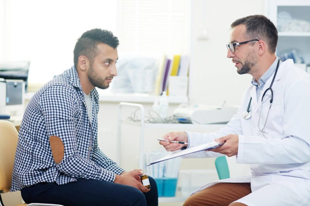 Vista lateral de homem jovem adulto conversando com um médico dentro de um consultório.