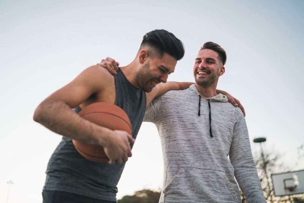 Vista de frente de dois homens rindo e praticando esportes enquanto um deles segura uma bola de basquete nas mãos.